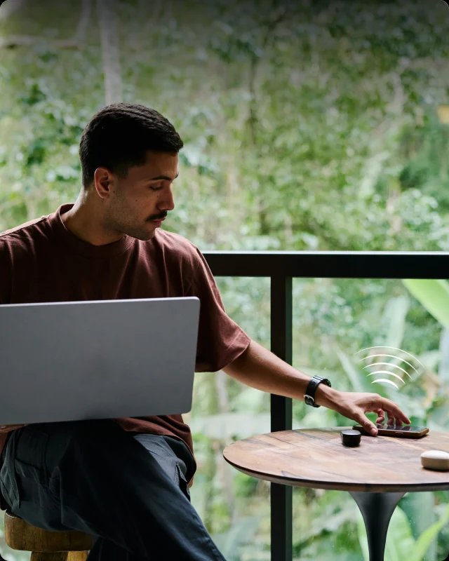 A man on a balcony with a laptop on his knees, unlocking his door from the phone on the table